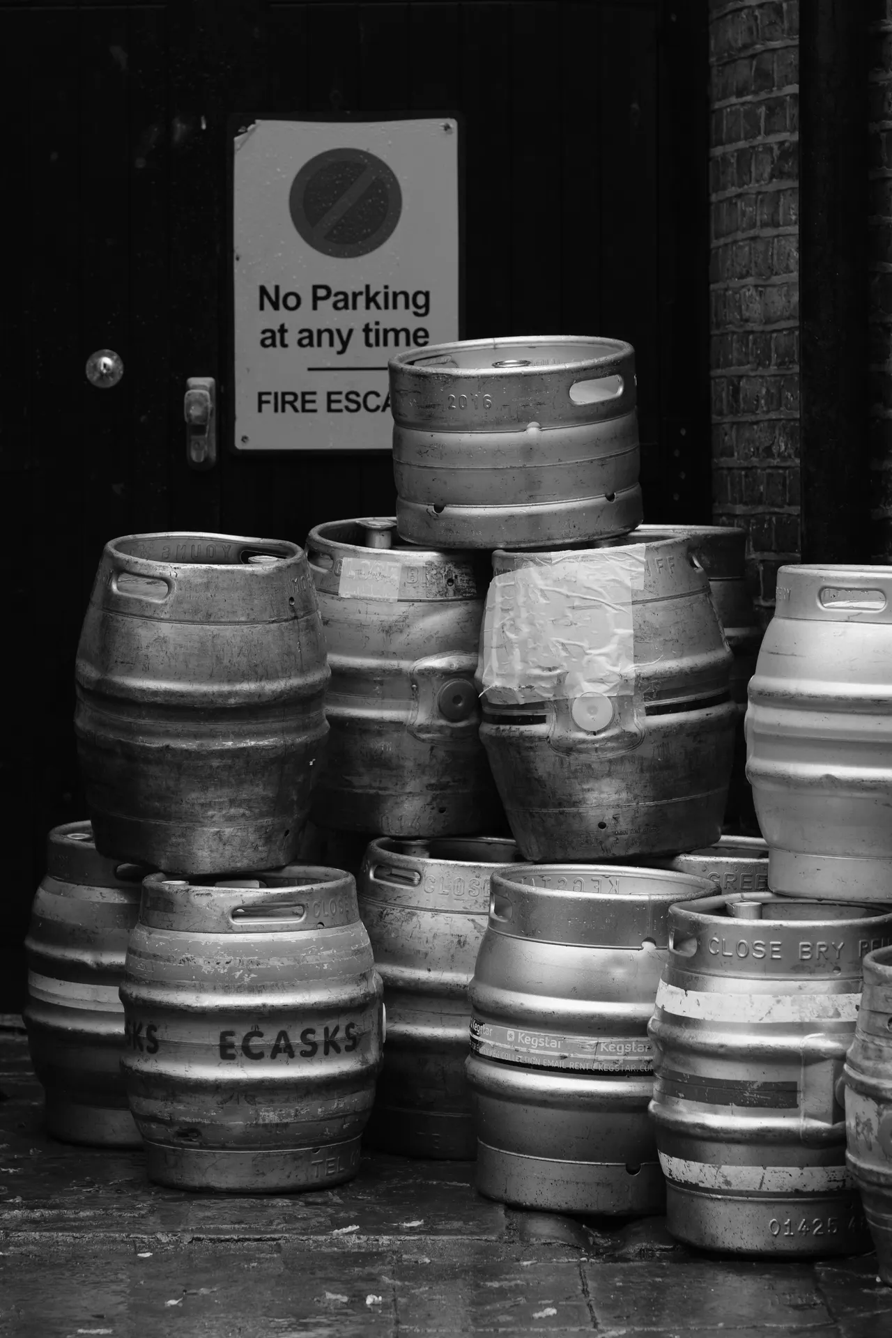 Metal beer kegs stacked by a "No Parking / Fire Escape" door, black and white