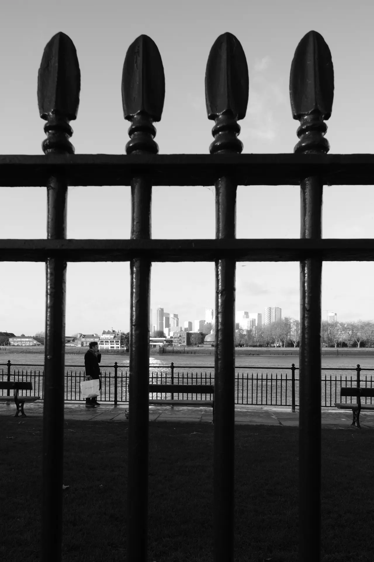 Canary Wharf towers behind iron railings, a passer-by along the Thames, black and white