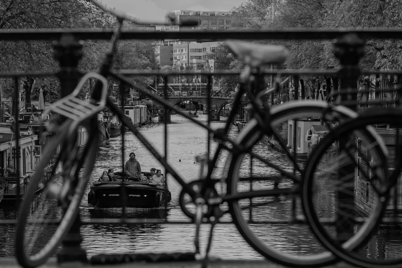 Vélo en silhouette sur la rambarde d'un pont, bateau de promenade passant dessous, noir et blanc