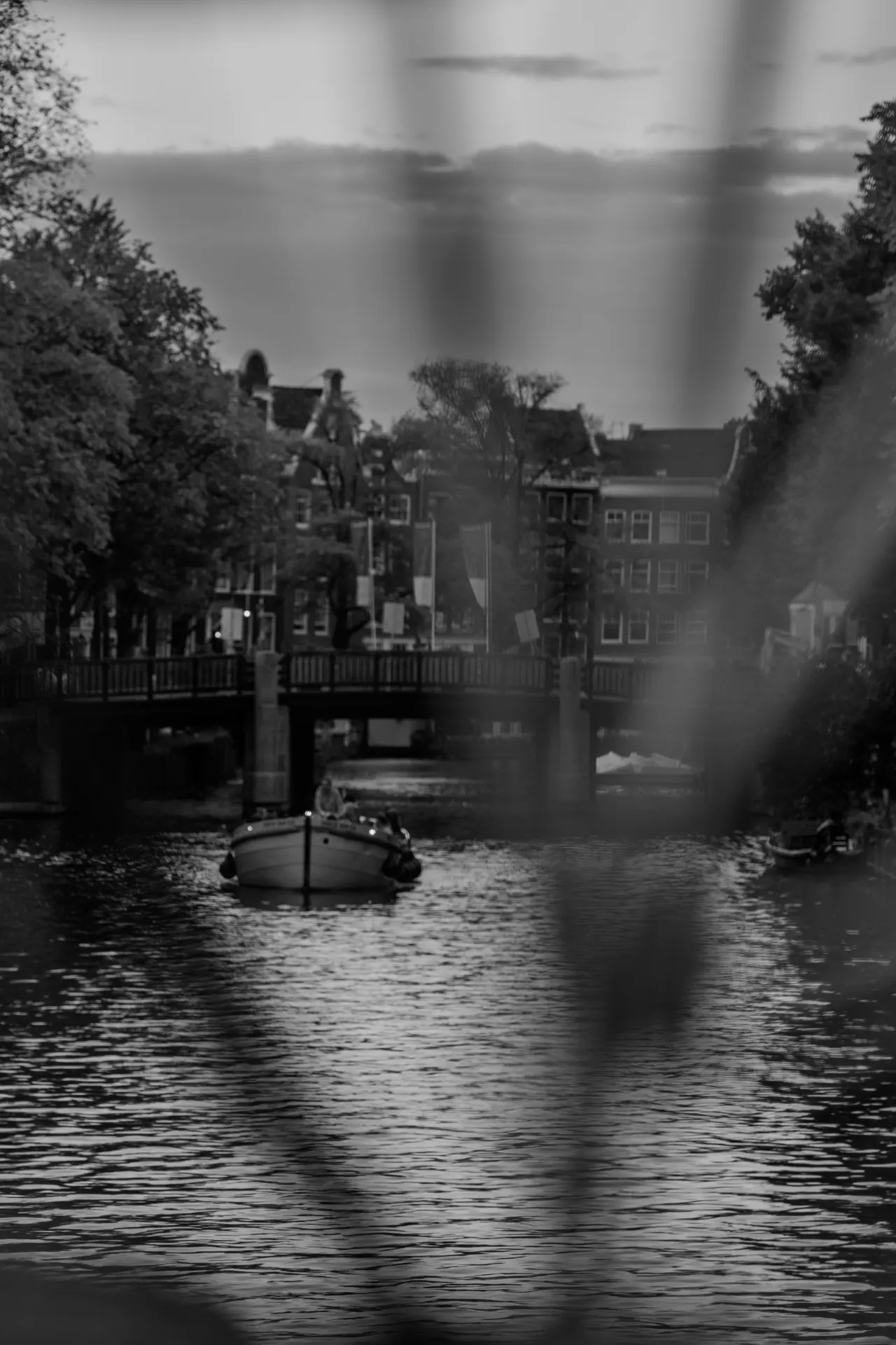 Bateau remontant le canal vers un pont, vu à travers les barreaux flous d'une rambarde, noir et blanc