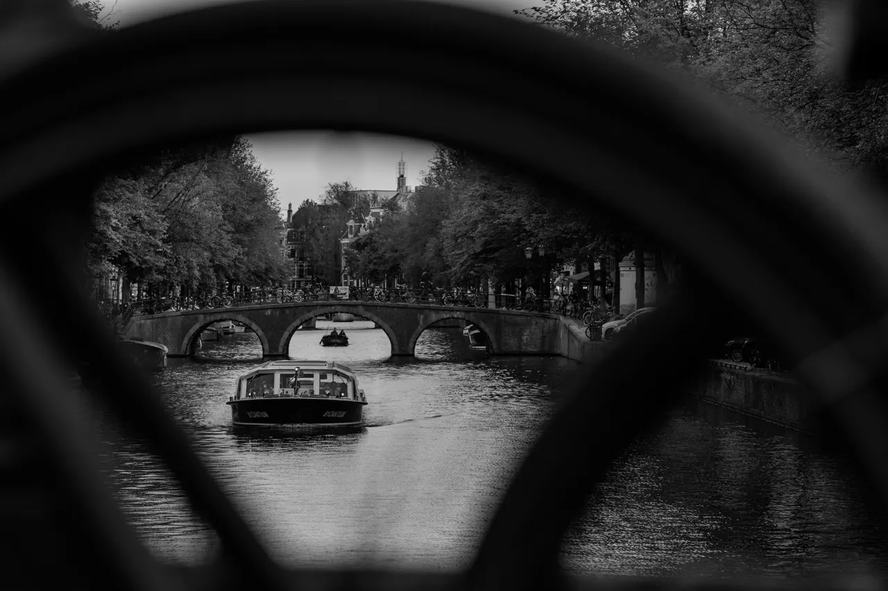 Bateau passant sous un pont à trois arches, cadré dans la roue d'un vélo, noir et blanc