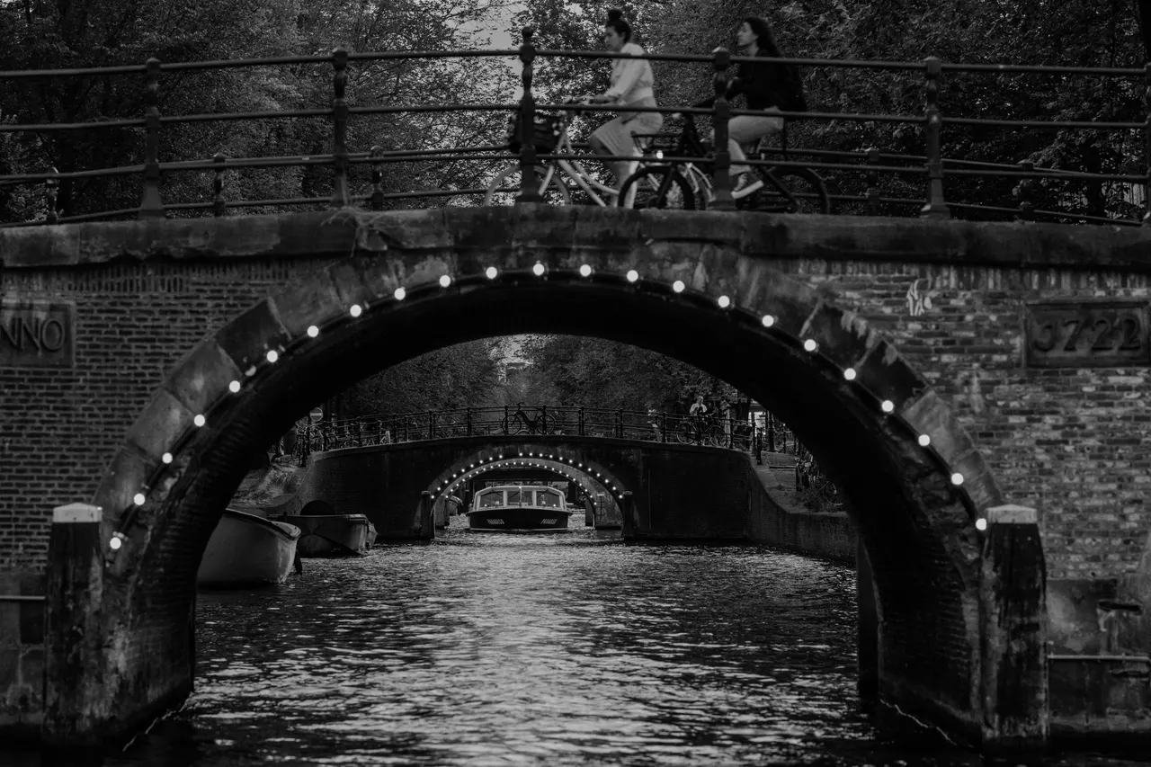Deux arches de pont éclairées alignées, cyclistes traversant au-dessus, bateau passant au fond, noir et blanc