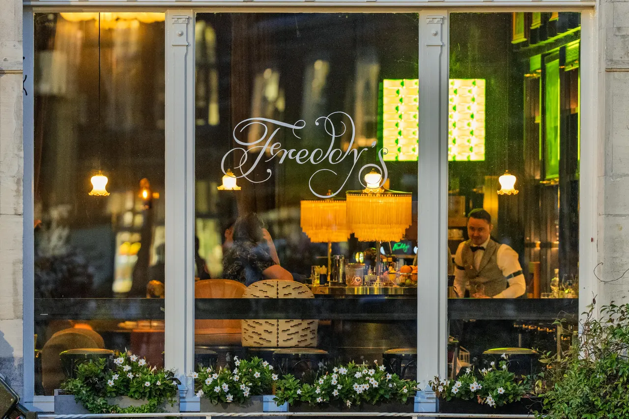 Barman derrière la vitrine éclairée d'un bar, lampes à franges et néon vert, vue depuis la rue
