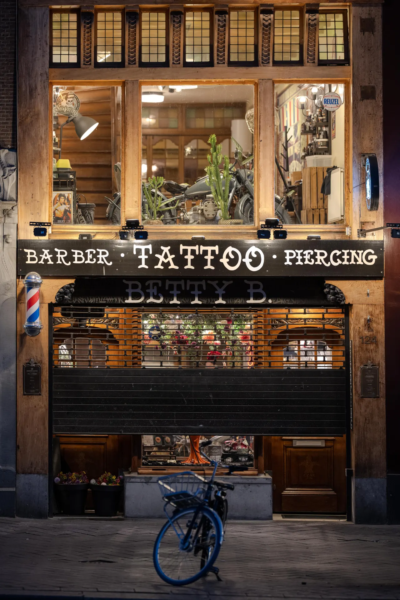 Devanture d'un barbier-tatoueur la nuit, rideau à demi baissé, vélo bleu garé devant