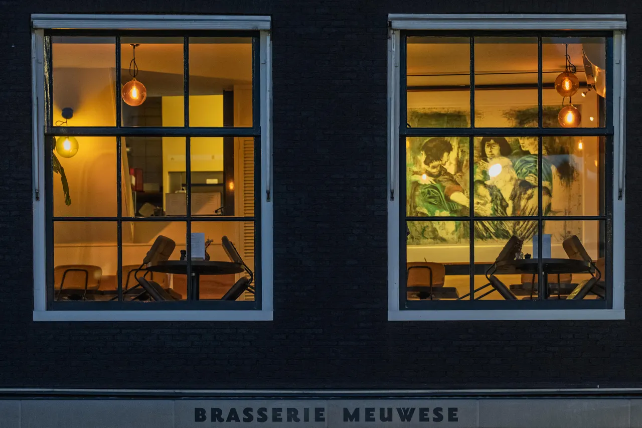 Deux fenêtres éclairées d'une brasserie dans une façade de brique noire, tables dressées et grande toile peinte