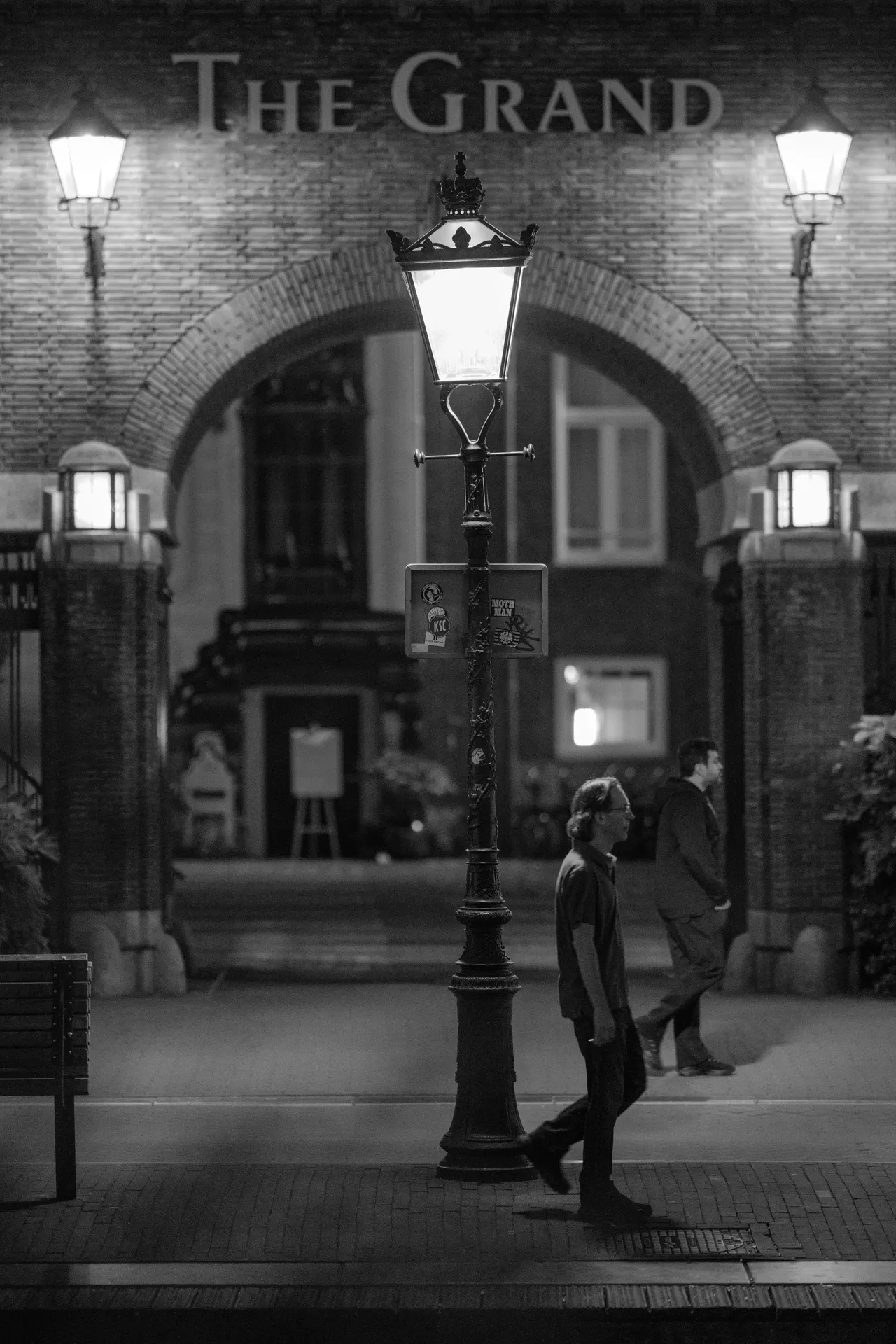 Réverbère ancien devant l'entrée voûtée de l'hôtel The Grand, deux passants marchant en sens inverse, noir et blanc