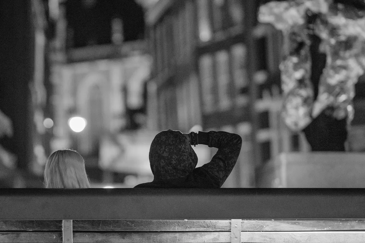 Deux personnes de dos sur un banc au bord du canal, un bras replié derrière la tête, façades floues au fond, noir et blanc