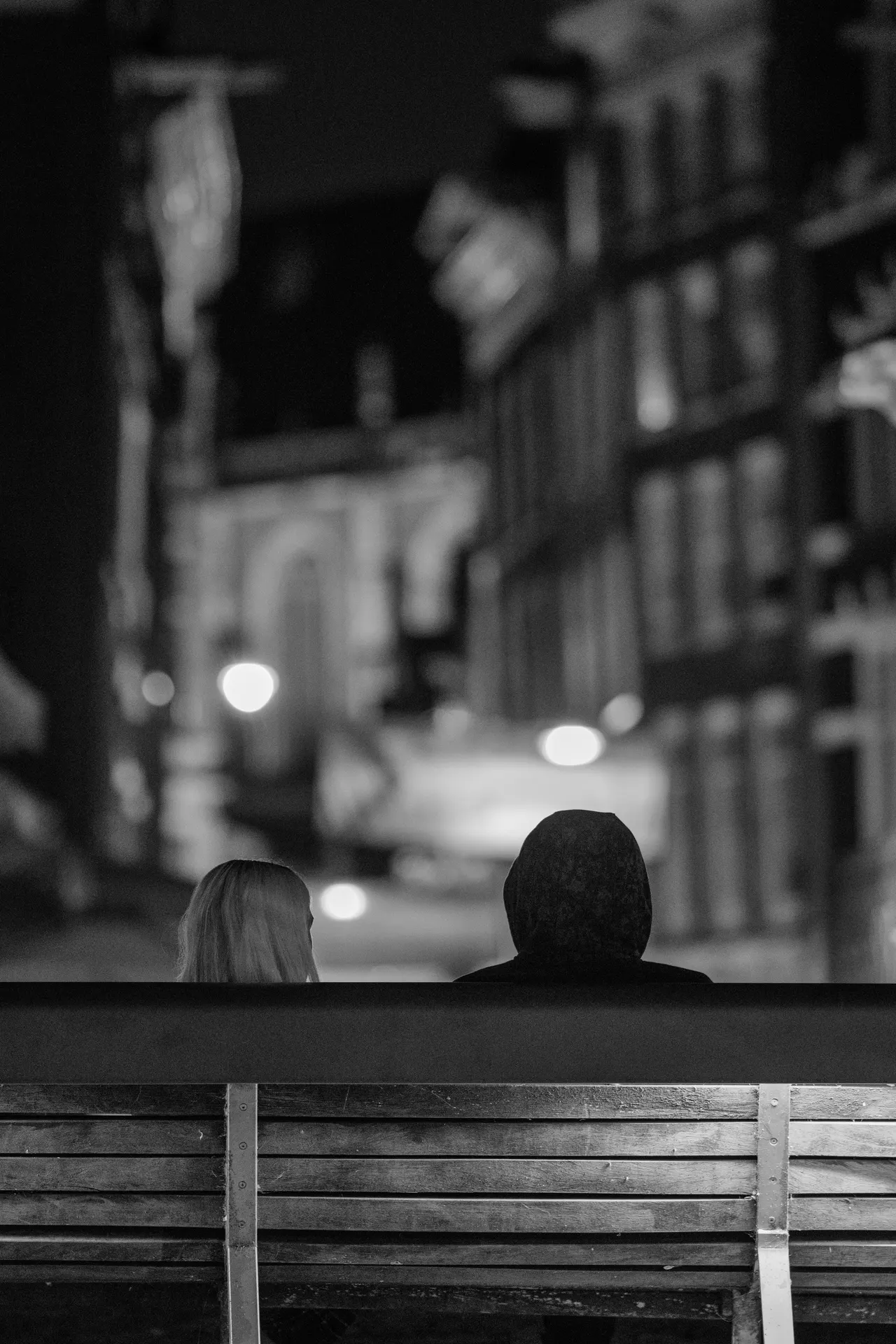 Deux silhouettes assises côte à côte sur un banc face au canal, lumières de rue floues au loin, noir et blanc
