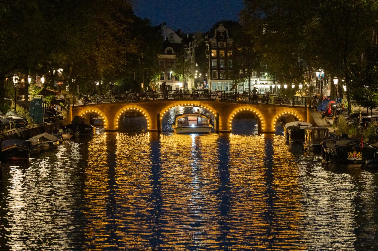 Pont du Reguliersgracht aux arches soulignées d'ampoules, bateau passant dessous, reflets dorés sur l'eau