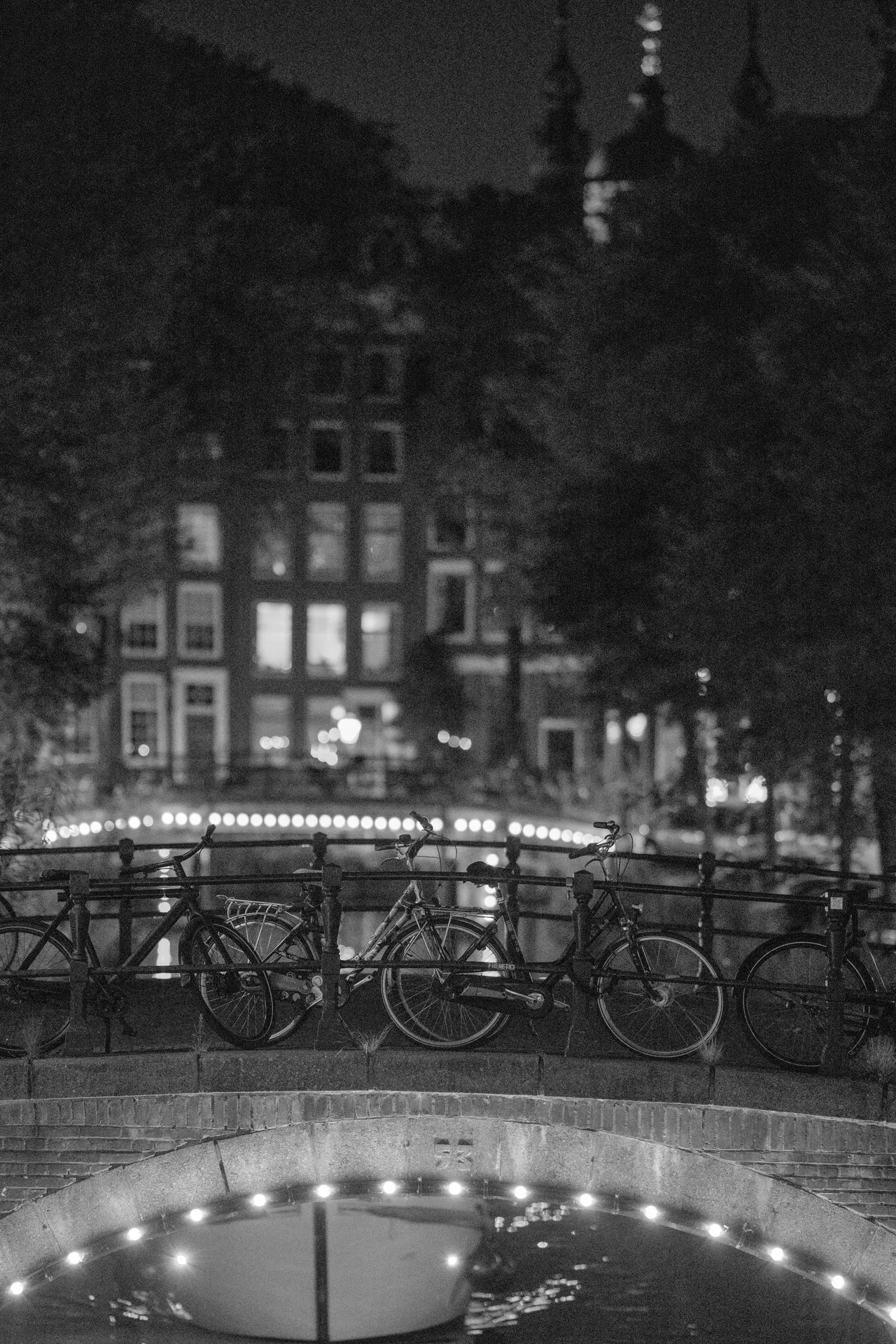 Vélos alignés contre la rambarde d'un pont de canal, arche éclairée d'ampoules au premier plan, noir et blanc