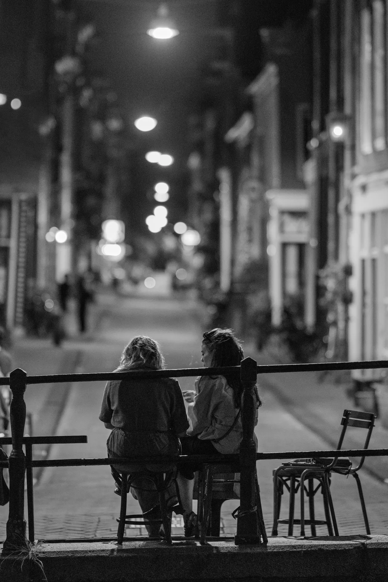 Deux femmes assises à une table de terrasse au bord d'un canal, rue déserte et enfilade de lampadaires au fond, noir et blanc