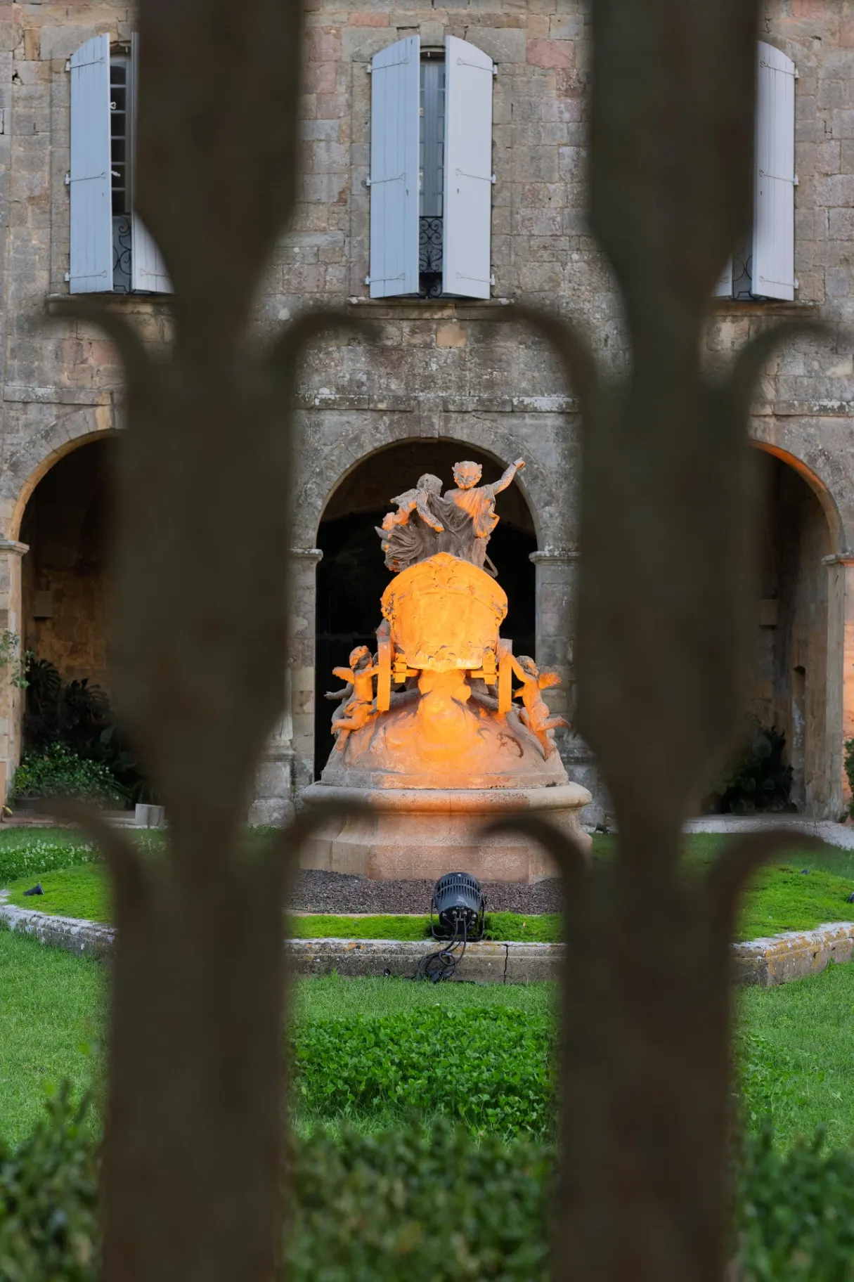 Baroque statue lit in orange behind a wrought-iron gate, courtyard of Fontfroide Abbey at night.
