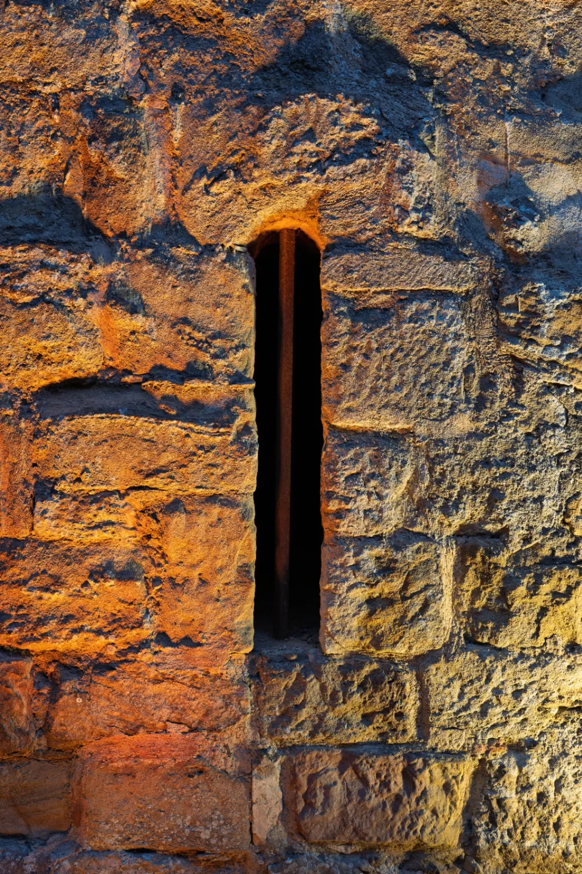 Vertical arrow slit in a sandstone wall split between warm orange and cold light, Fontfroide Abbey.